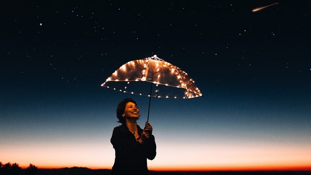 Foto de uma mulher sorridente, à noite, com um guarda-chuva com lâmpada acesas, simbolizando o benefício da educação empreendedora - criatividade